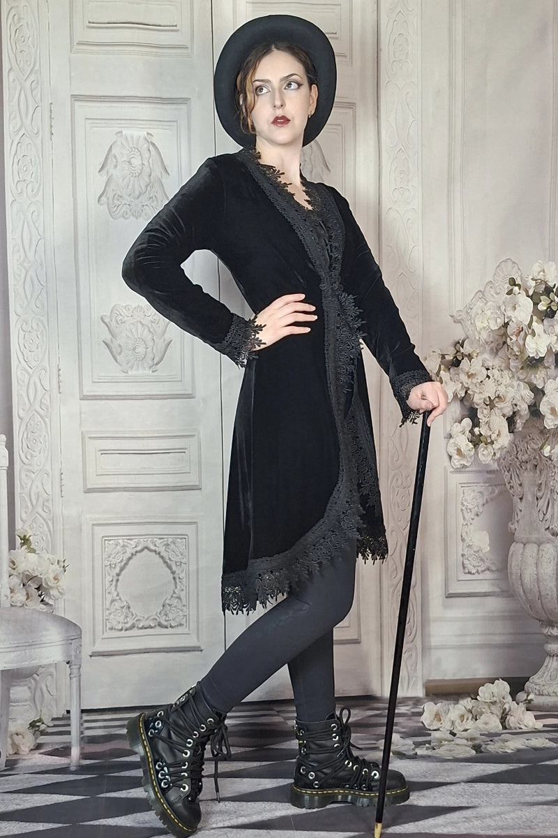 Woman in a black outfit with a hat and boots standing in an elegant room with decorative walls and flowers.