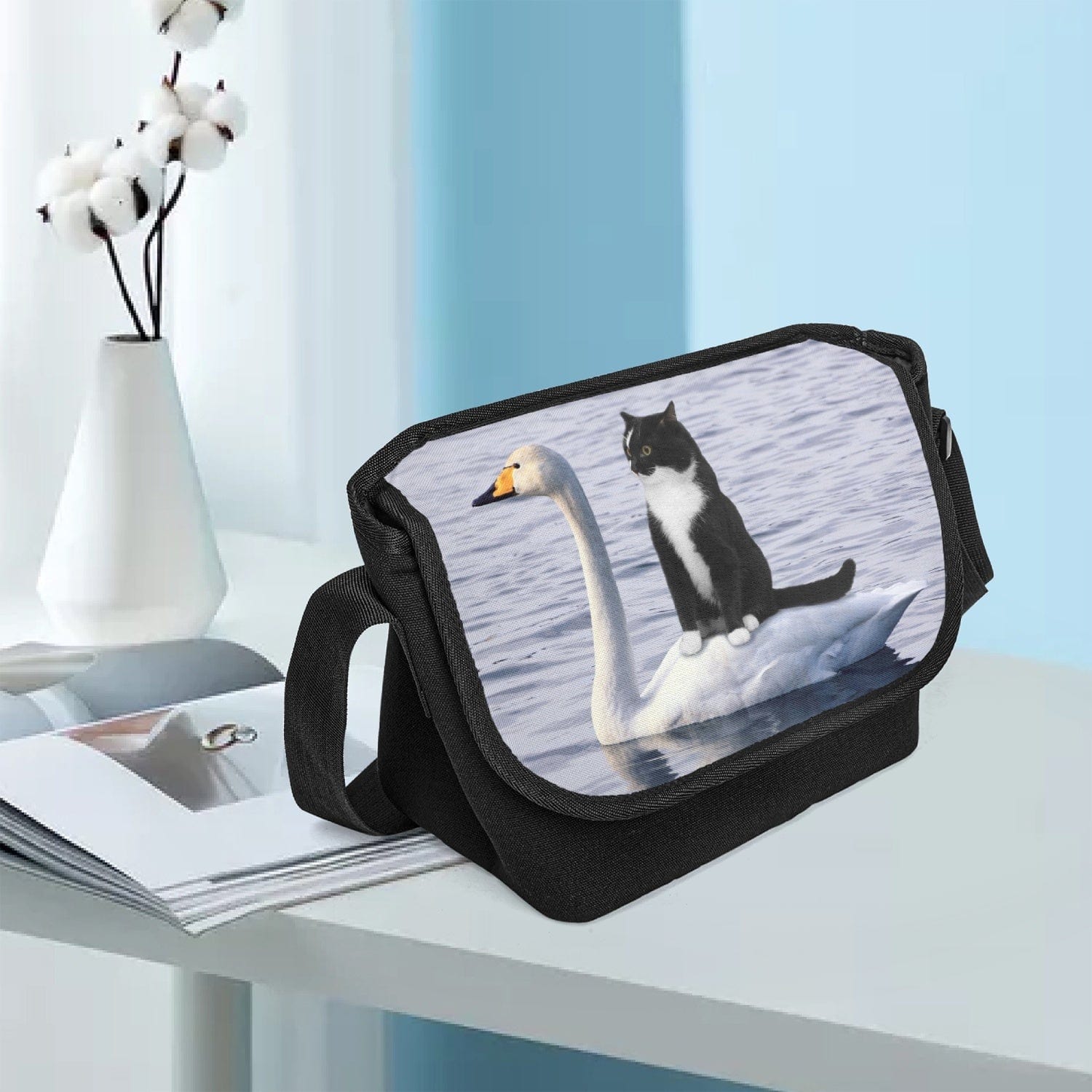 a tuxedo kitten rides on a white swan across a pond, a photographic print on a black canvas messenger bag sitting in a phd's office