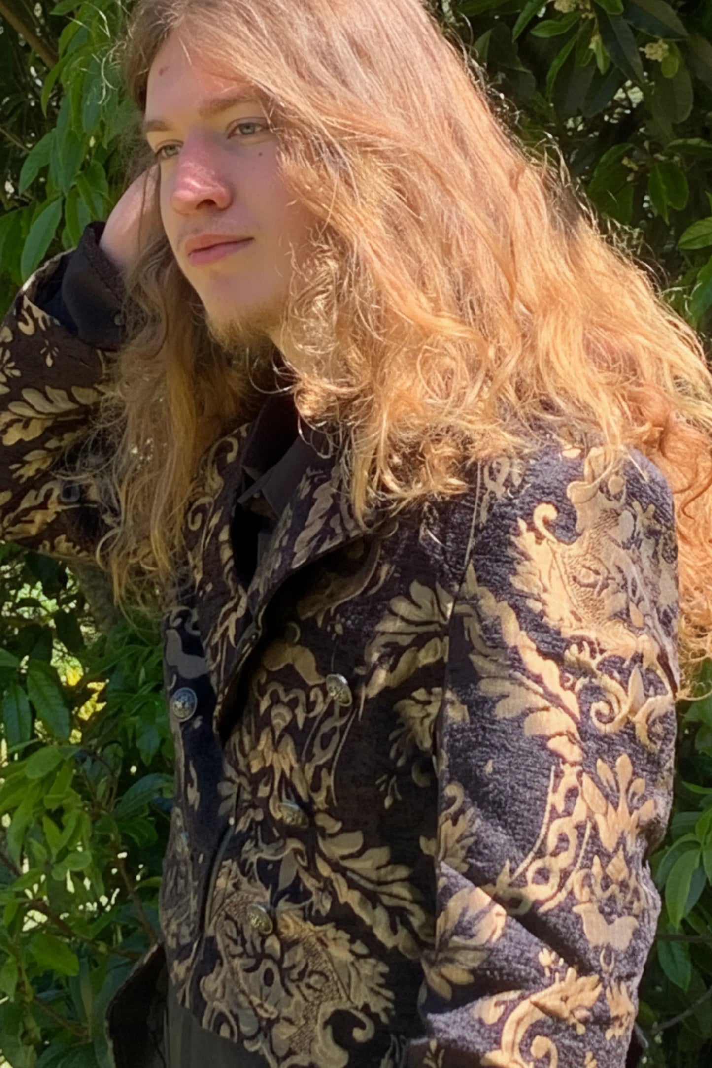 Person wearing a patterned coat standing outdoors with greenery in the background