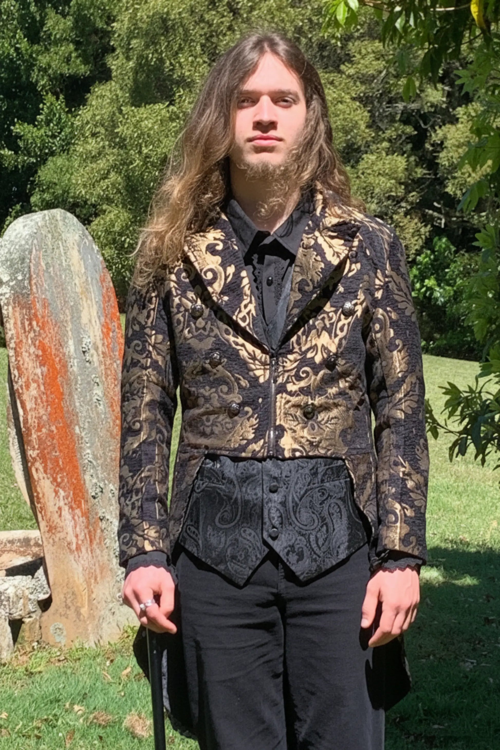 Person wearing a patterned jacket and black pants standing outdoors with trees in the background