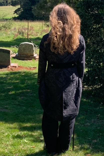 Person wearing a dark coat standing in a grassy area with trees and a gravestone in the background