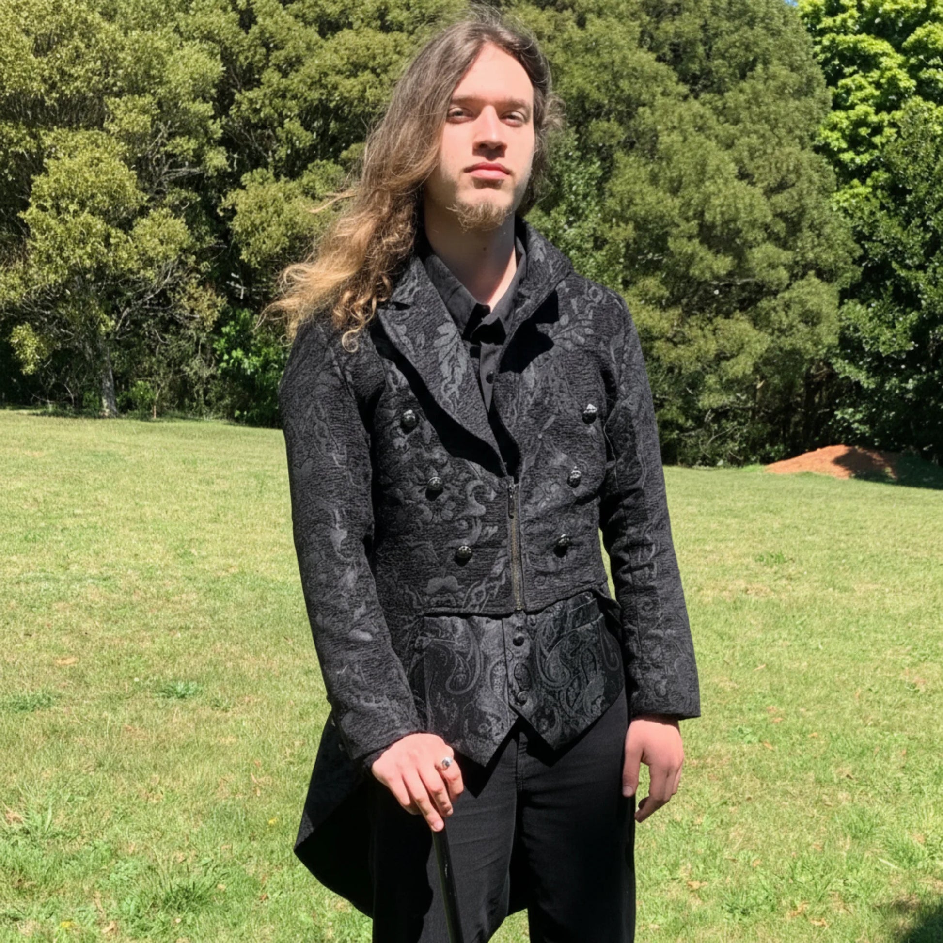 Man wearing a dark patterned coat standing in a grassy field with trees in the background