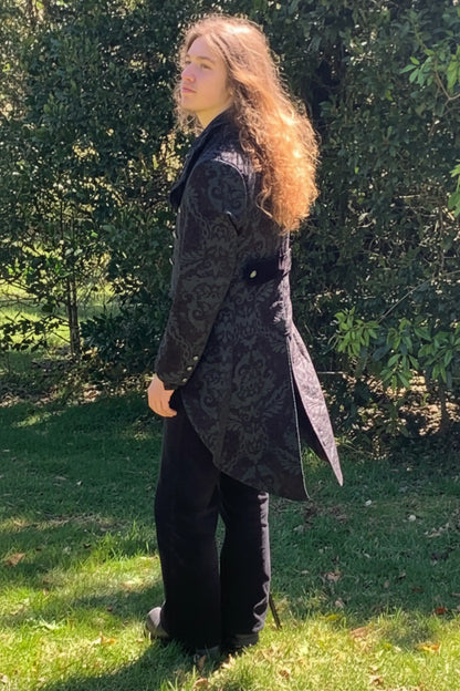 Person wearing a long dark coat standing outdoors with greenery in the background