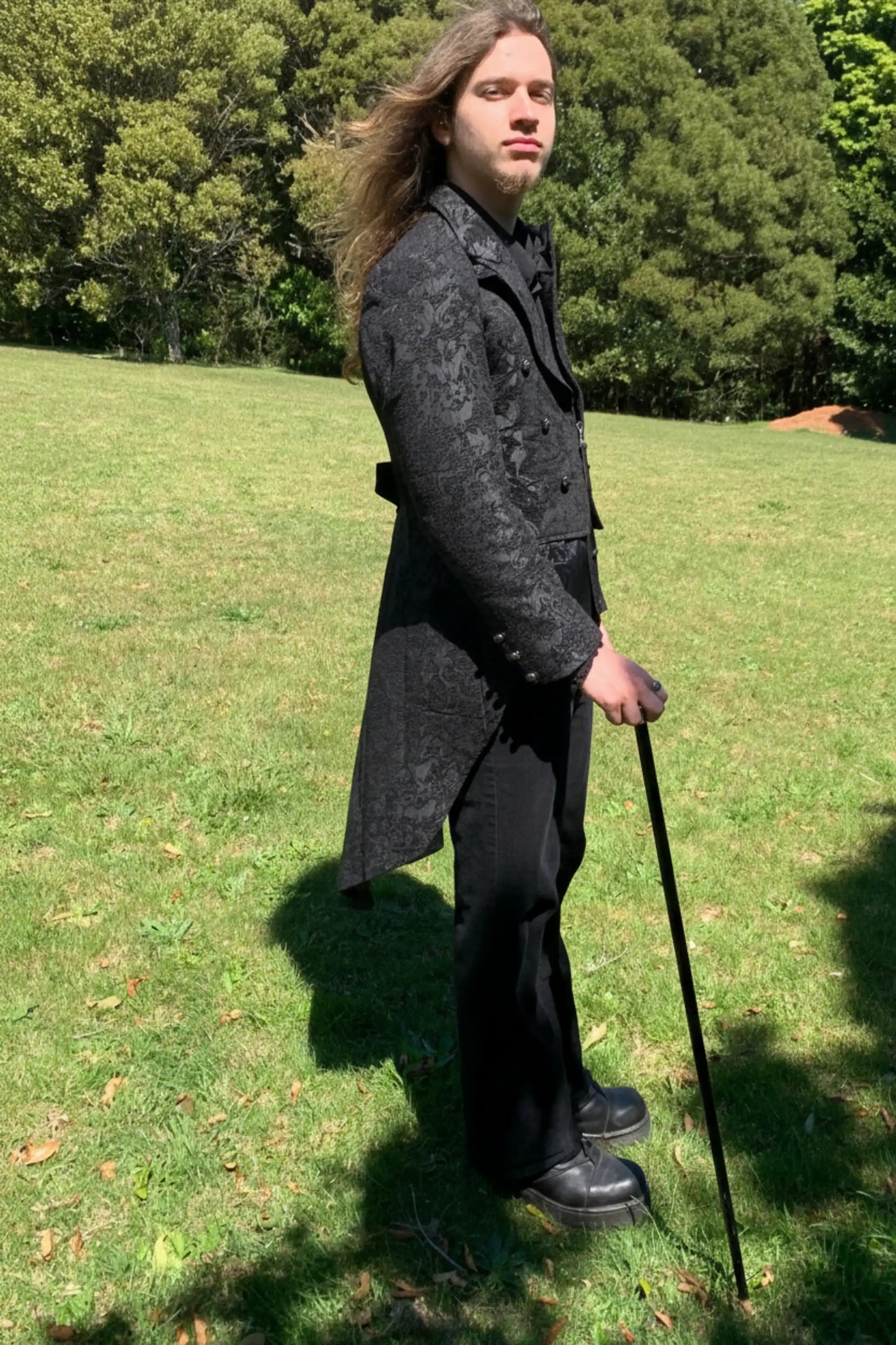 Person in a black formal outfit with a long coat and cane standing on grass with trees in the background