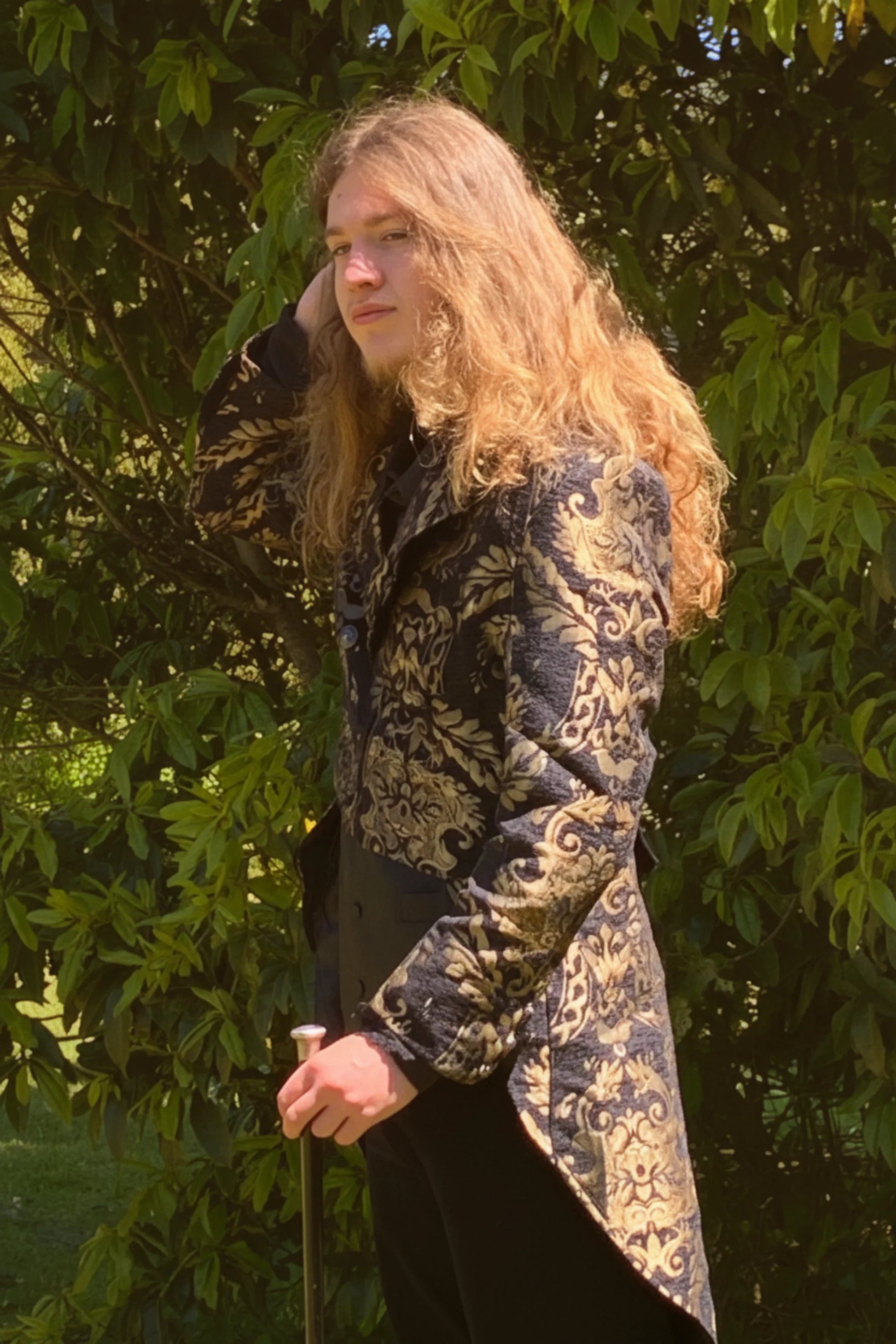 Gothic male wearing a baroque tail coat standing outdoors with greenery in the background