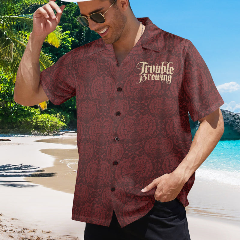 Man wearing a 'Trouble Brewing' shirt on a beach with palm trees and blue sky.