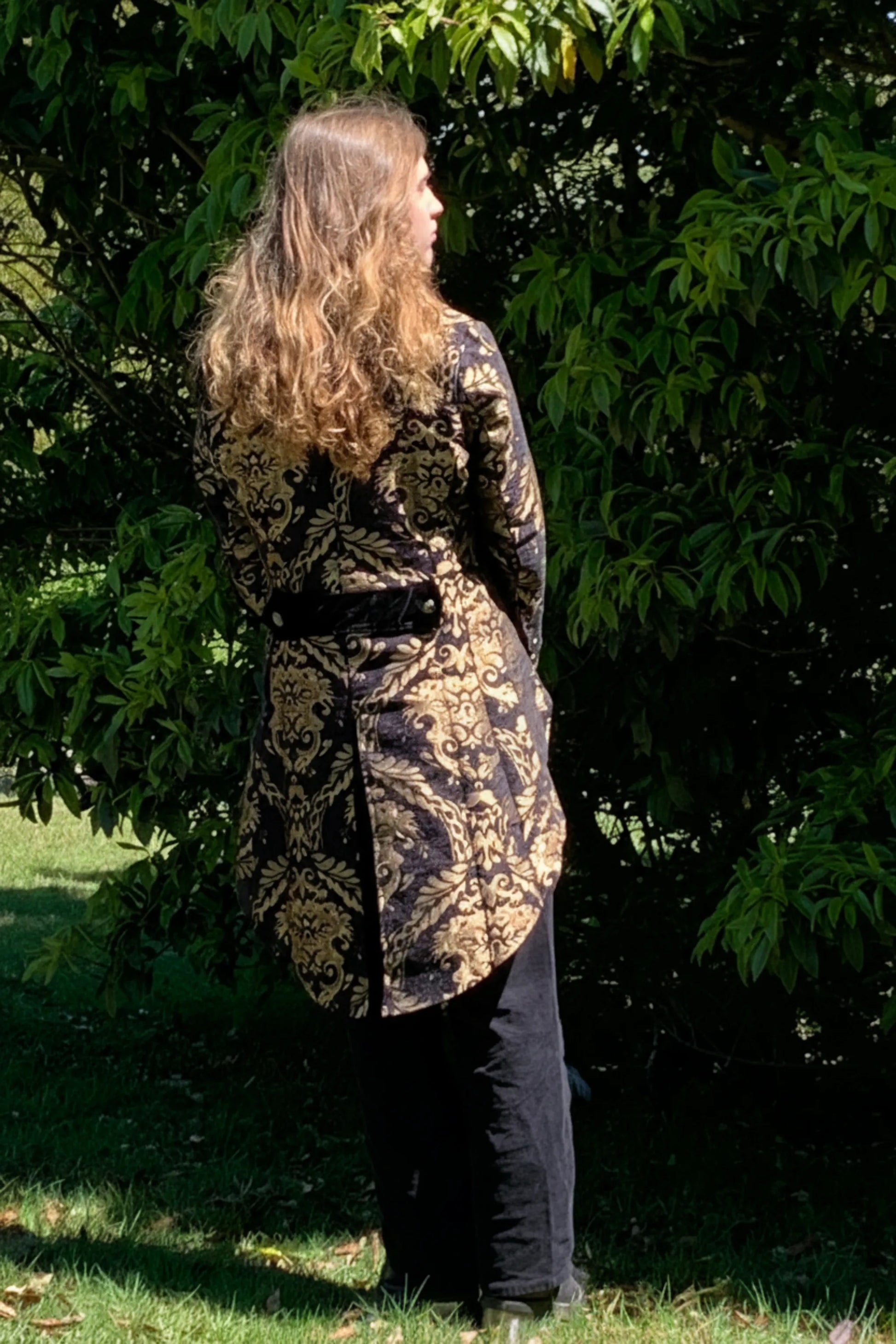 Gothic male with long hair wearing a gold and black brocade tail coat standing in a garden