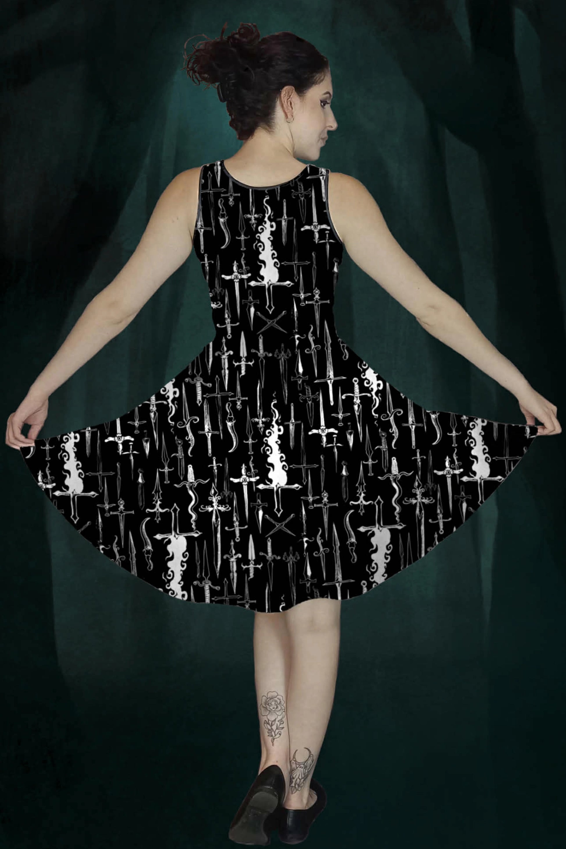 Woman wearing a black dress with white dagger & flames patterns against a dark green enchanted forest background, back view