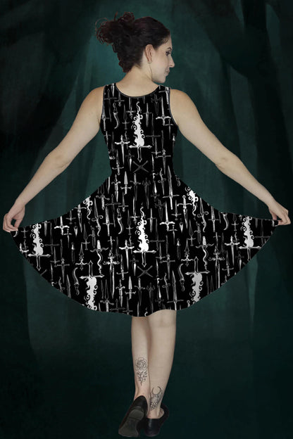 Woman wearing a black dress with white dagger & flames patterns against a dark green enchanted forest background, back view