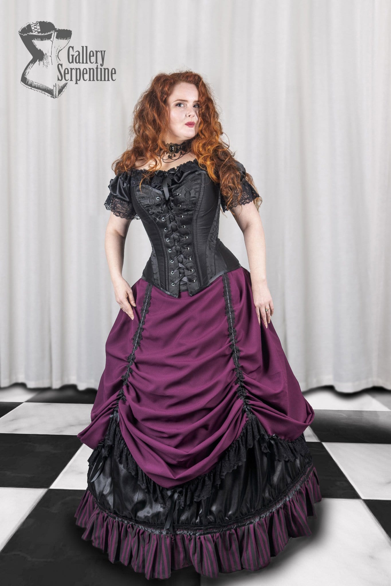 front view of a red headed model wearing a black steel boned over bust corset and burgundy victorian skirt with a hoop underneath it