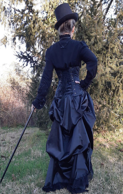 full back view of victorian woman wearing a corset over the gothic victorian black sateen lace trimmed high low bustle skirt with adjustable waist sizing made in Australia for Gallery Serpentine